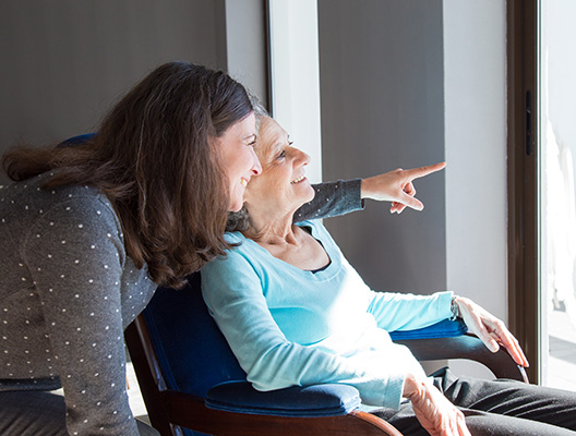 Positive mother and daughter enjoying dramatic view out of window. Young health visitor showing to senior lady funny scene out of window. Family relations concept